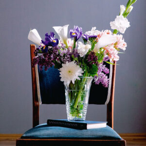 side-view-bouquet-white-color-calla-lilies-with-dark-purple-iris-lilac-white-gladiolus-flowers-glass-vase-standing-book-chair-grey-wall-background
